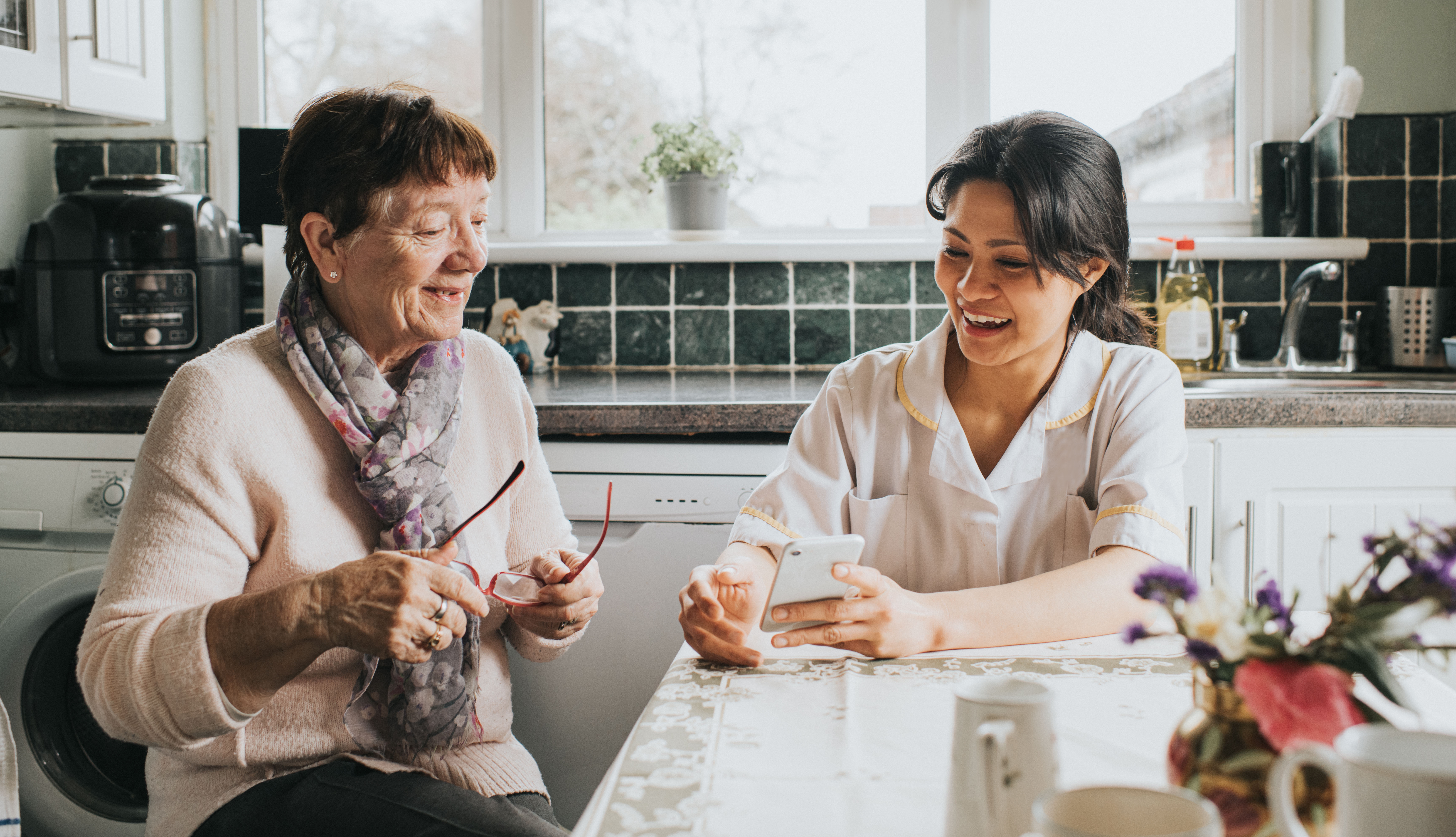 Una cuidadora sonriente se sienta junto a una mujer mayor en una mesa de cocina. Ella ayuda a la mujer a usar un teléfono inteligente para realizar un pedido en línea.