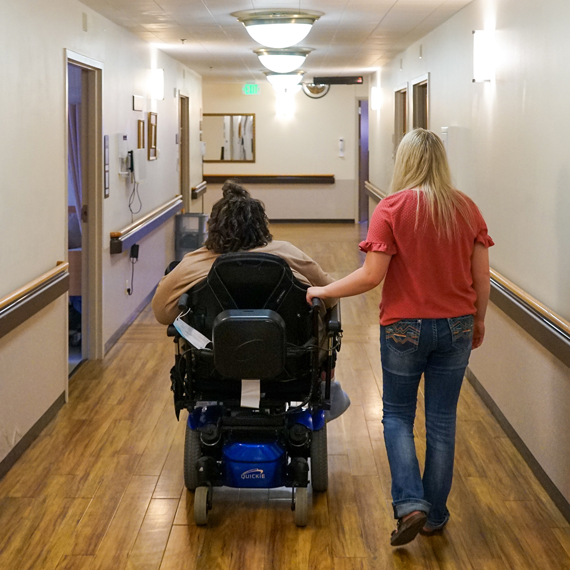 nursing home resident in wheelchair accompanied by activities aide walk down a hall
