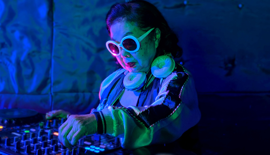 item 5 of Gallery image - Sumiko, a well know DJ in Tokyo, standing behind a mixing board with headphones around her neck and sunglasses on