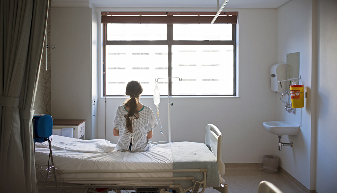 A female patient in a hospital gown sitting on a hospital bed looking out the window