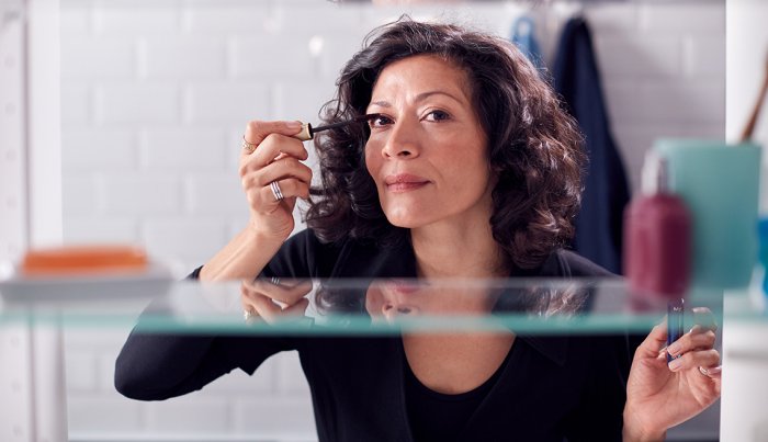 A view through a bathroom cabinet of a woman applying mascara