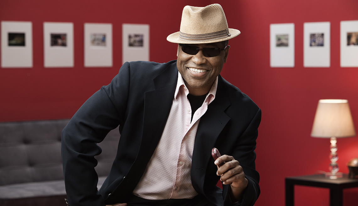 A man smiling while wearing a hat sunglasses suit jacket and collared shirt