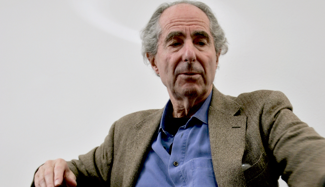 item 16 of Gallery image - Philip Roth leans back in a chair at a table wearing a suit coat.