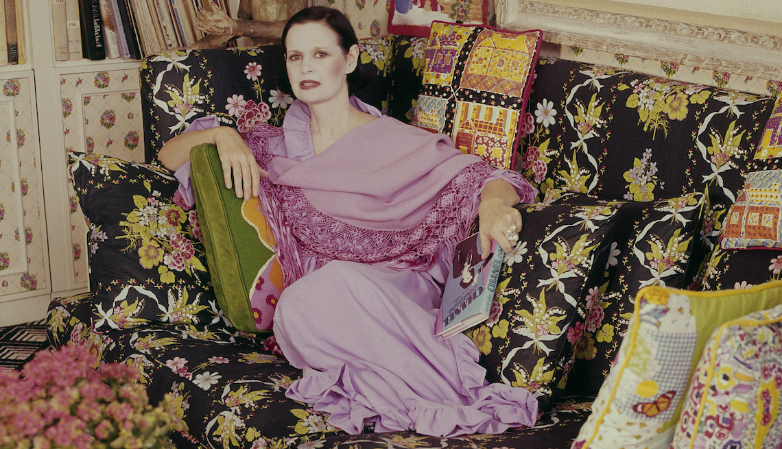 item 18 of Gallery image - Gloria Vanderbilt culred up on a couch with flowered printed pillows she designed.