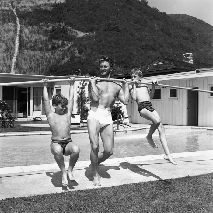 black and white photo from the fifties showing actor kirk douglas lifting his sons michael and joel up in his backyard using a pole