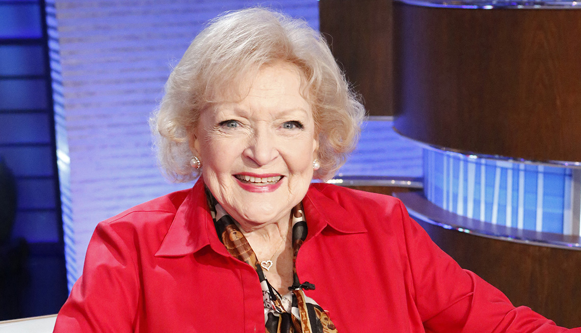 Betty White in a red shirt smiling on the set of the game show To Tell the Truth