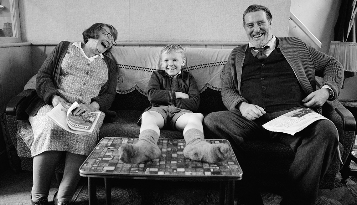 Judi Dench, Jude Hill and Ciaran Hinds laughing and smiling together on a couch in the film Belfast