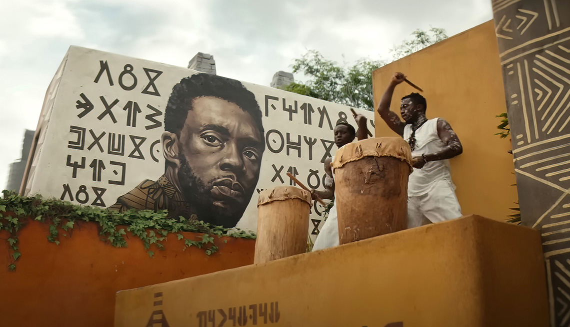 A mural of Chadwick Boseman painted on a wall with two people drumming nearby in the film Black Panther: Wakanda Forever