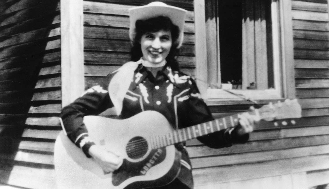 Loretta Lynn holds her acoustic guitar as she poses for a portrait wearing a cowboy hat, a scarf and western shirt outside a log cabin in circa 1960.