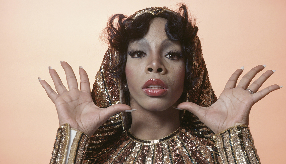 item 14 of Gallery image - Headshot of Donna Summer, US singer-songwriter, posing in a studio portrait, with her hands raised, open-palmed, either side of her head, wearing gold-sequinned cuffs and collar, 1976.
