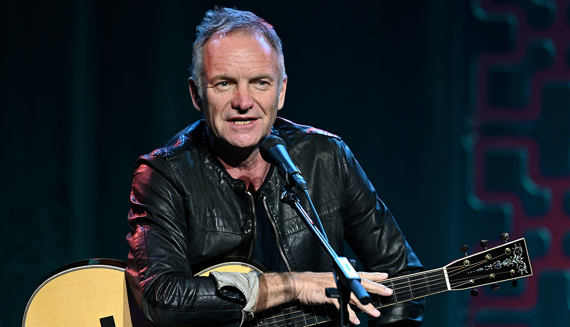 Sting on stage with an acoustic guitar talking into a microphone