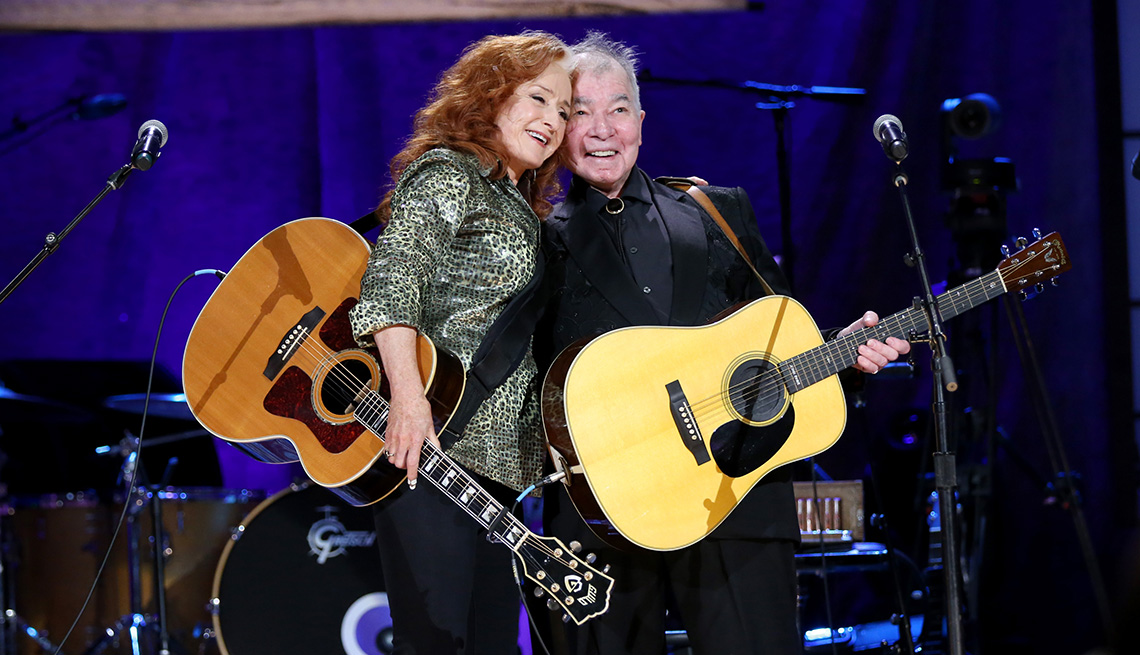 Bonnie Raitt and John Prine embrace on stage while holding their guitars during the 2019 Americana Honors and Awards