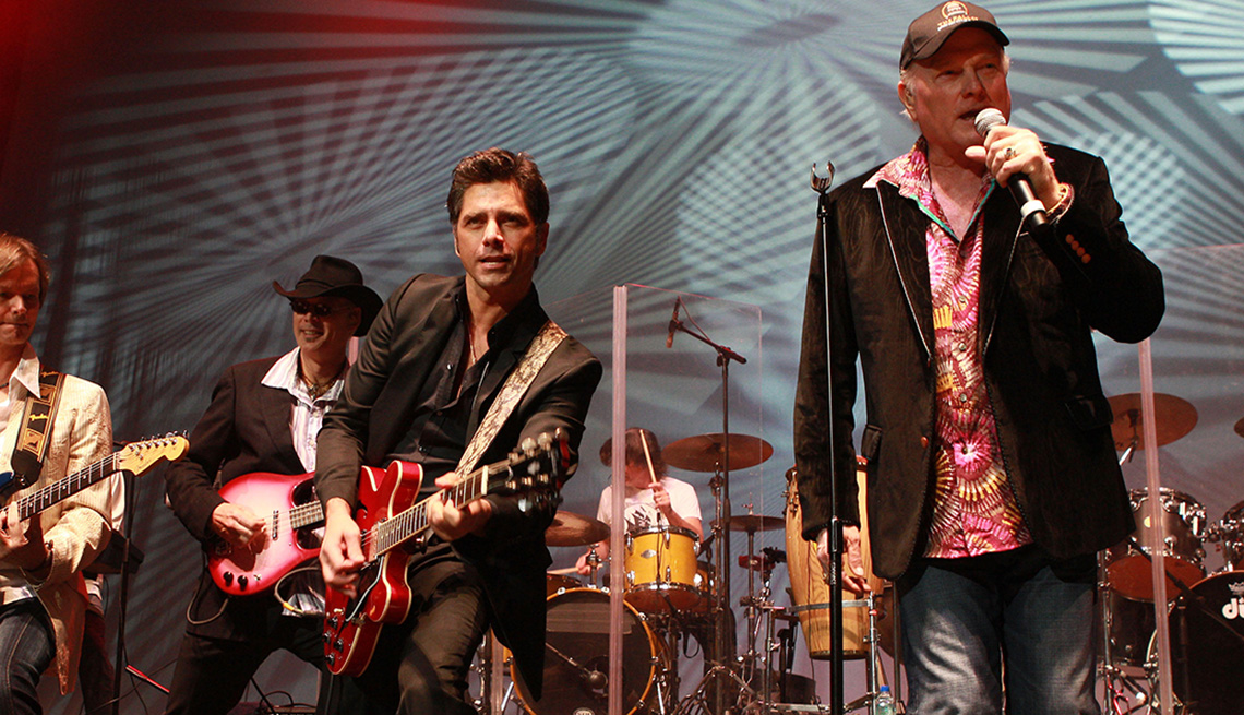 John Stamos holding a guitar and performing on stage with the Beach Boys.