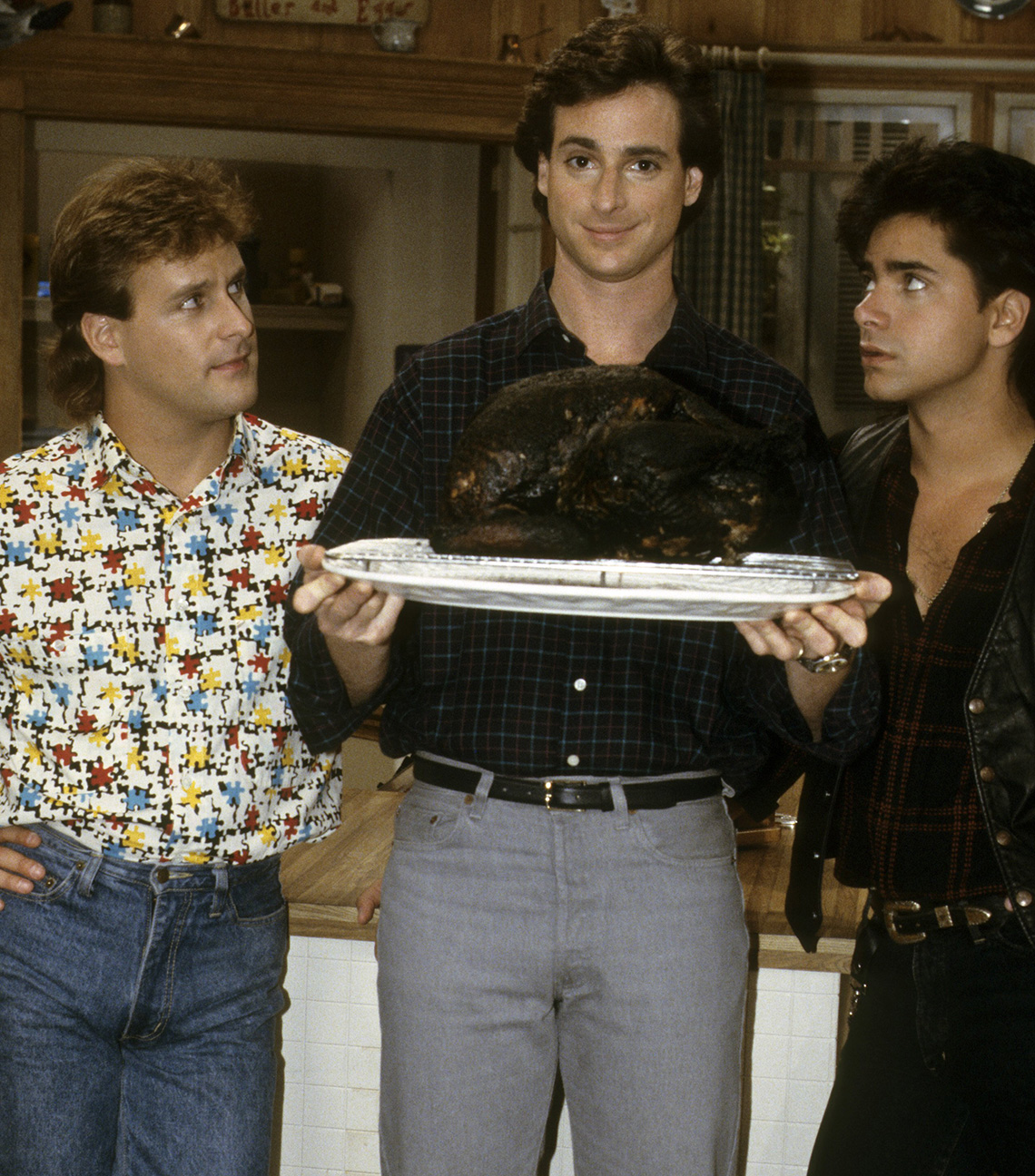 Bob Saget holding a completely burnt turkey with Dave Coulier and John Stamos looking and standing beside him in a scene from Full House