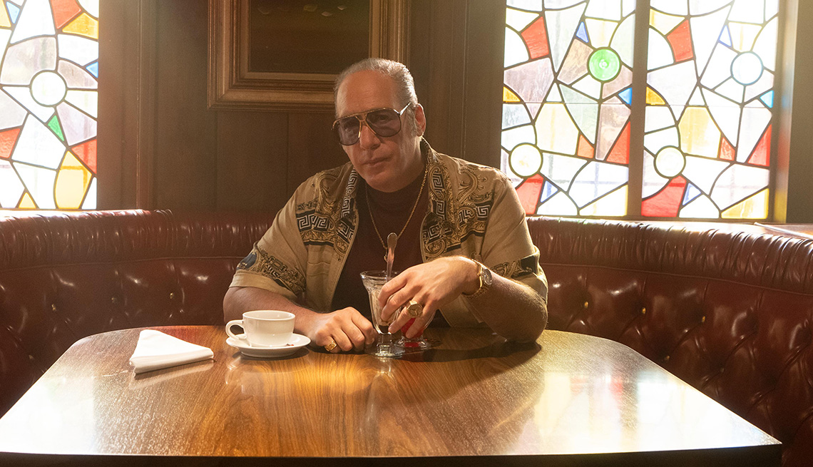 Andrew Dice Clay sitting at a table with a coffee cup and a glass drink on the table in the Hulu series Pam and Tommy