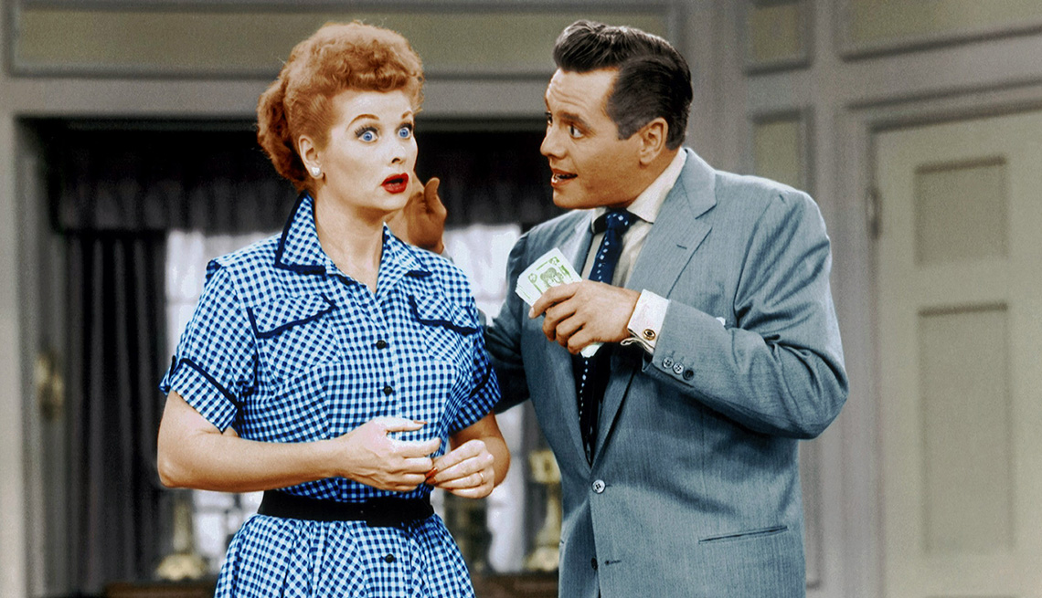 Lucille Ball with a shocked look on her face standing next to Desi Arnaz holding cash in his hand while talking to her in the television show I Love Lucy