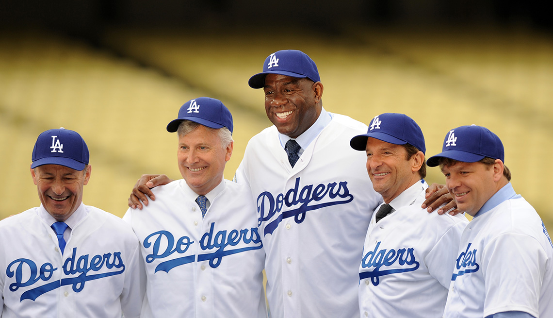 Stan Kasten, Mark Walter, Magic Johnson, Peter Guber, and Todd Boehly stand side by side posing for a photo at a press conference introducing them as the new owners of the Los Angeles Dodgers