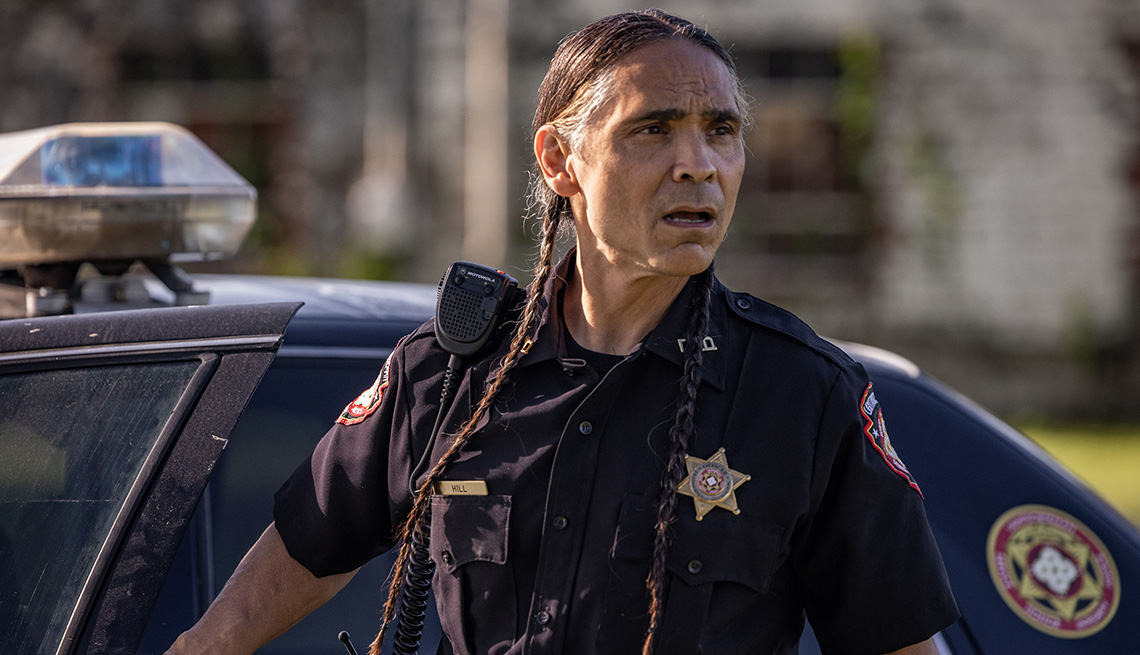 Zahn McClarnon wearing his officer uniform in front of a police vehicle in the television series Reservation Dogs