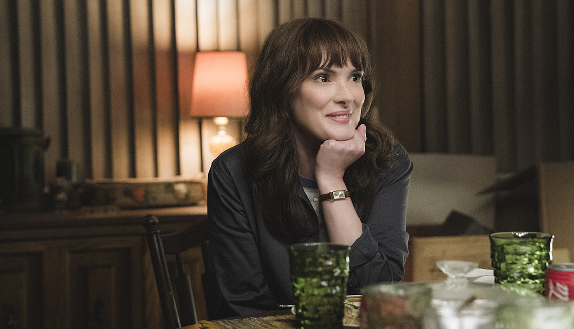 Winona Ryder sits at a dinner table with her chin being supported by her hand in a scene from Season 4 of Stranger Things