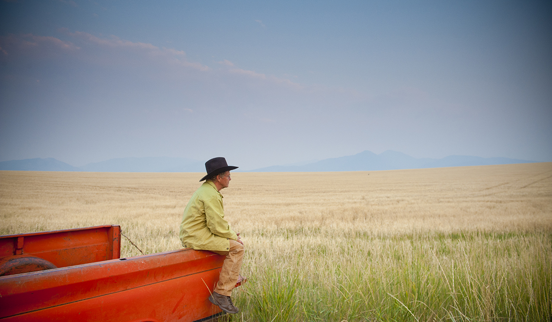 Farmer in brown hat sits on the edge of his red pickup truck in a wheat field.