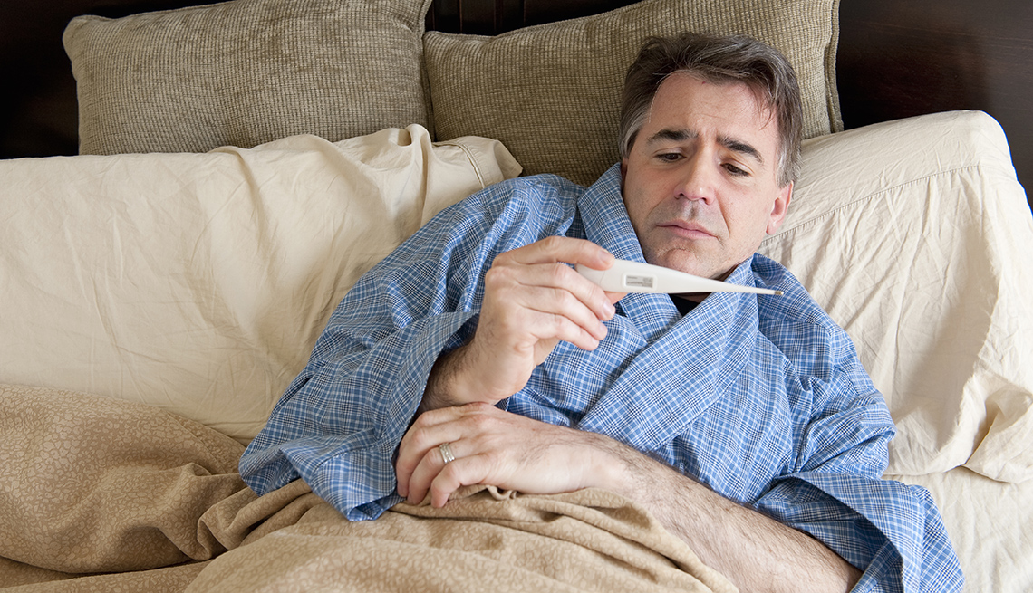 Mature man sick in bed, looking at thermometer. 