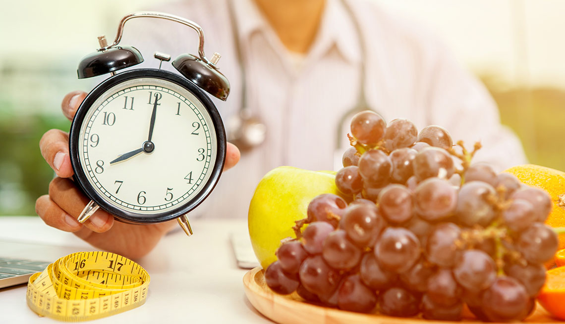 Una persona sostiene un reloj despertador y sobre una mesa hay uvas y una cinta de medir