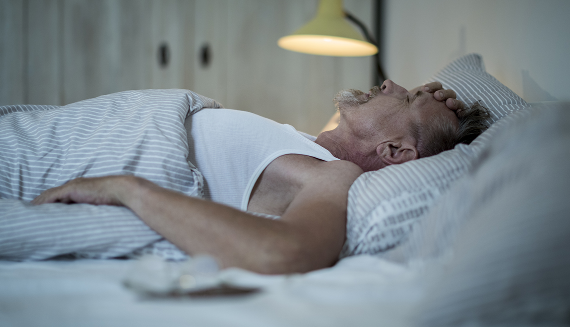 An older man is lying in bed holding his head.
