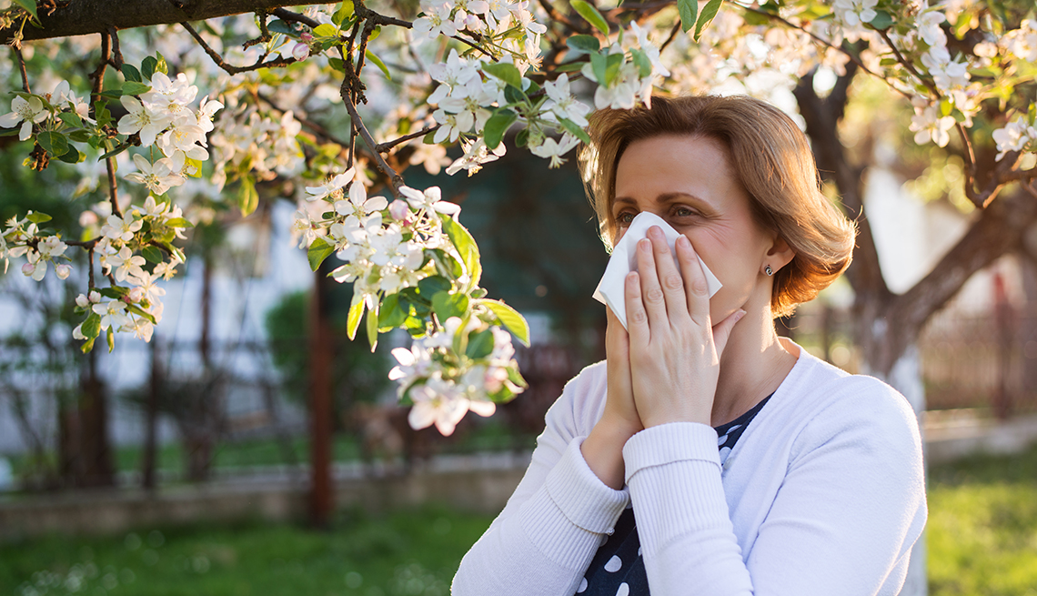 woman with pollen allergies standing outdoors under blossoming trees sneezing into a tissue