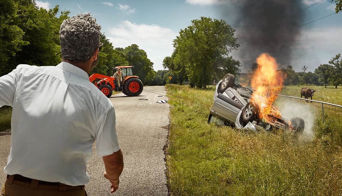 back of man running towards the scene of a vehicle accident on a country road with a car flipped over and on fire
