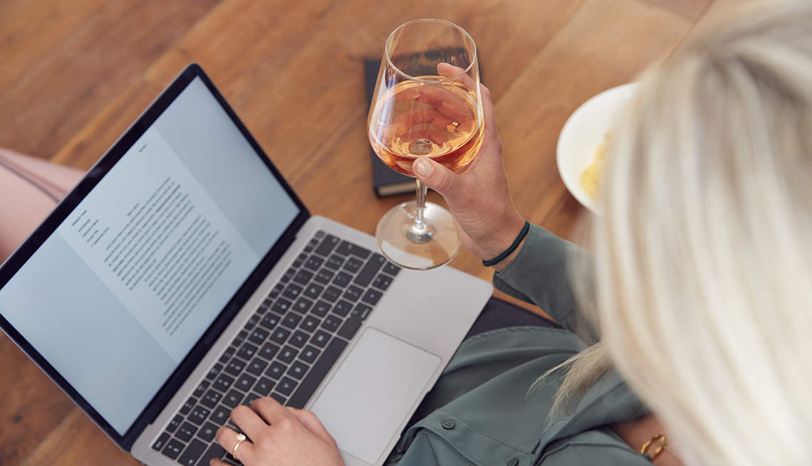 Una mujer trabaja desde su casa en una computadora, mientras sostiene una copa de vino