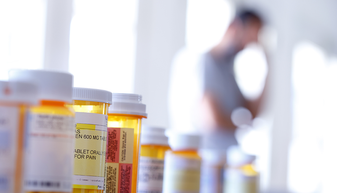 A large group of prescription medication bottles sit on a table as a man in the background stands with his hand on his head.