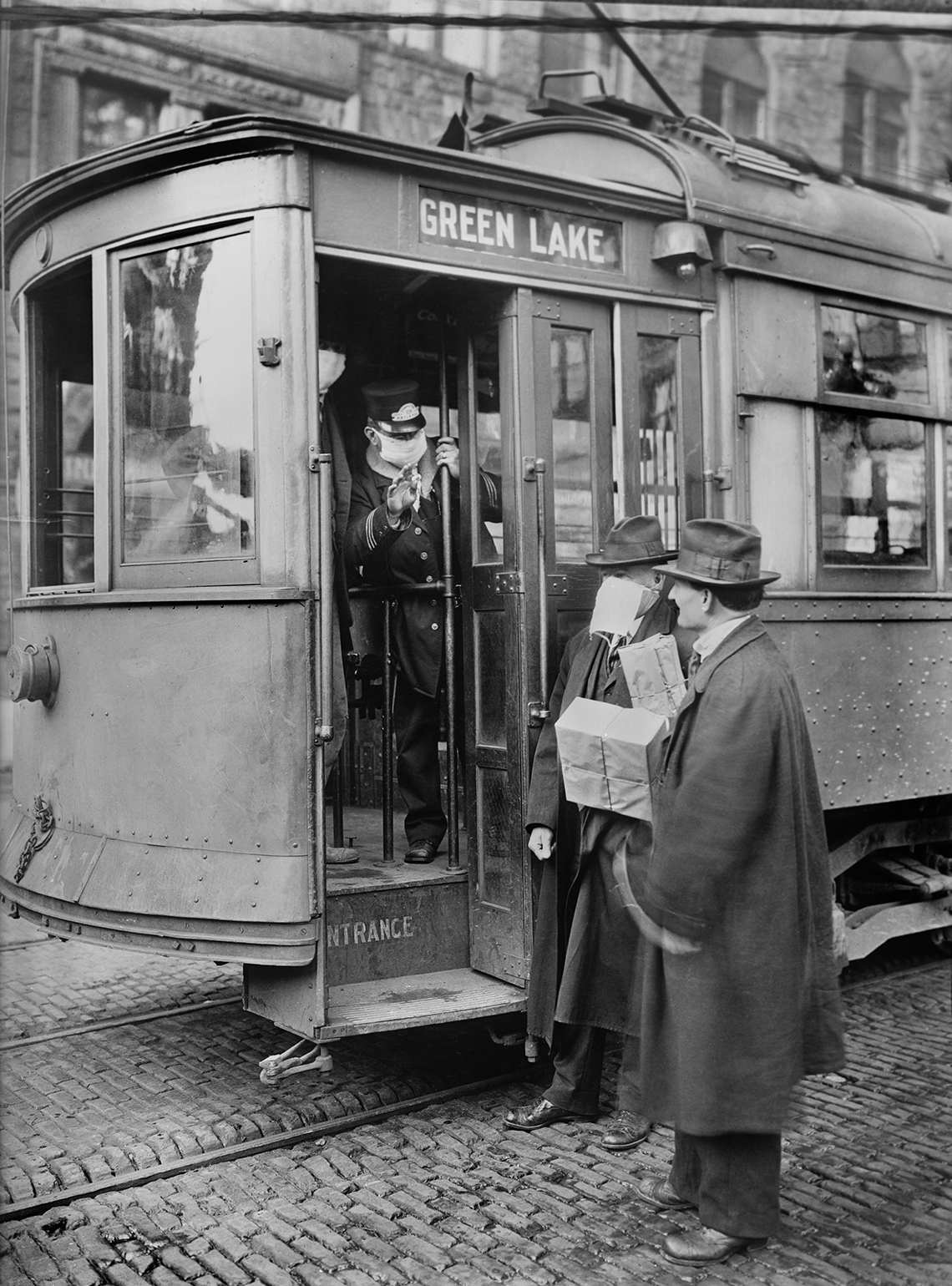 old photo of two men about to board a streetcar speaking with the driver who is wearing an anti-influenza face mask