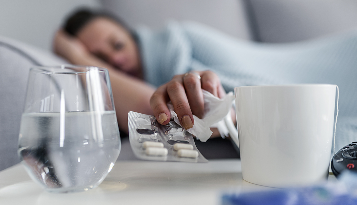 Una mujer acostada en su cama y en su mesita tiene agua, pastillas y una taza