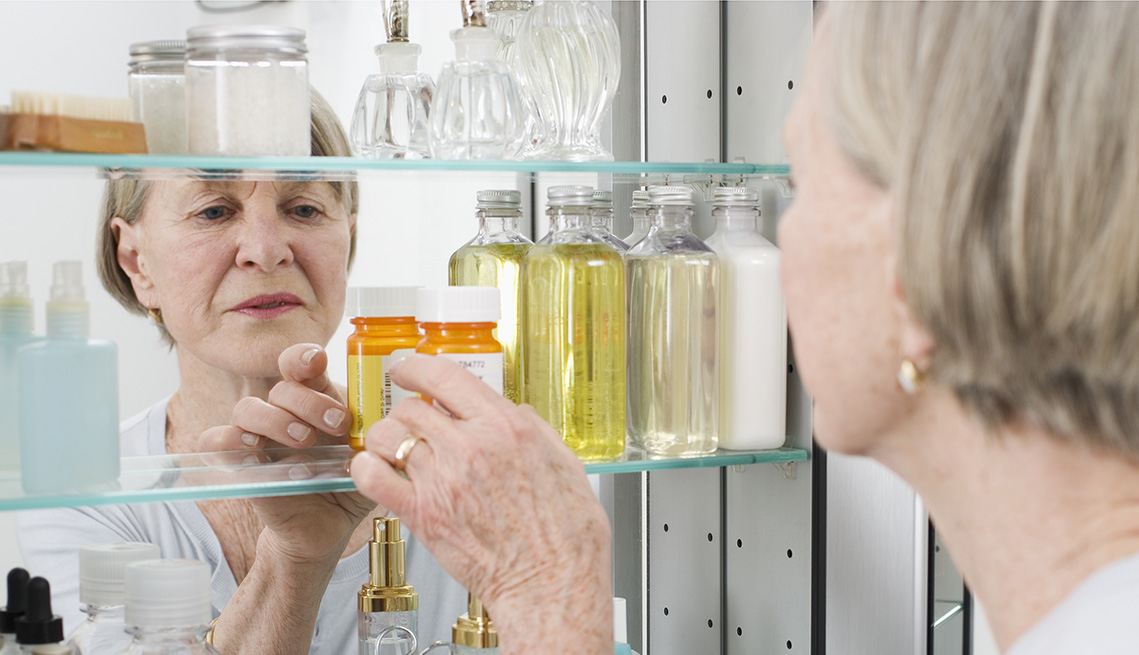 Mature woman getting pills out of a medicine cabinet. Her reflection in the mirror looks concerned. 