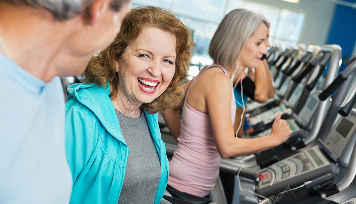 Mature woman working out in a gym, talking to a man who is also working out.  Another woman working out in the background.