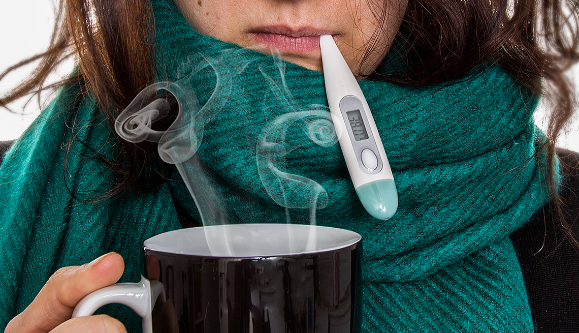 A woman with the flu, a green scarf is wrapped around her neck and a thermometer is in her mouth. She is holding a cup of hot tea.
