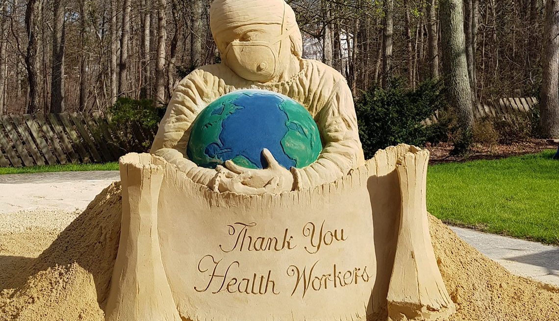 item 3 of Gallery image - A masked medical worker cradles the world in a six-ton sand sculpture built by artists John Gowdy and Laura Cimador-Gowdy at their home in Galloway, New Jersey.