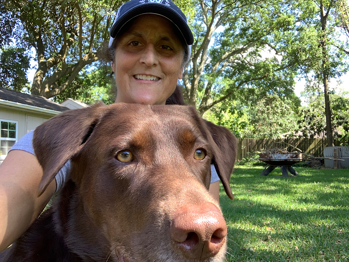 a woman in a baseball cap poses with her chocolate labrador retreiver dog in her home back yard