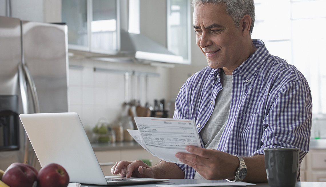 man at home holding a paper utility bill and paying it on his laptop