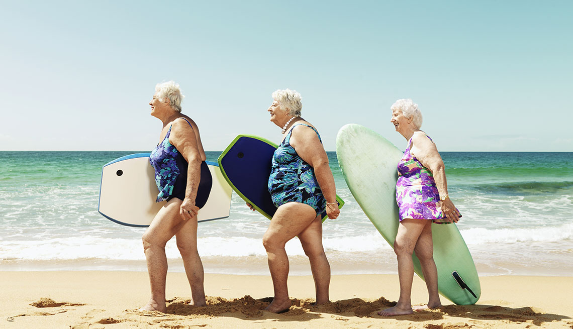 Tres mujeres mayores caminan por una playa cada una con una tabla para practicar el surf