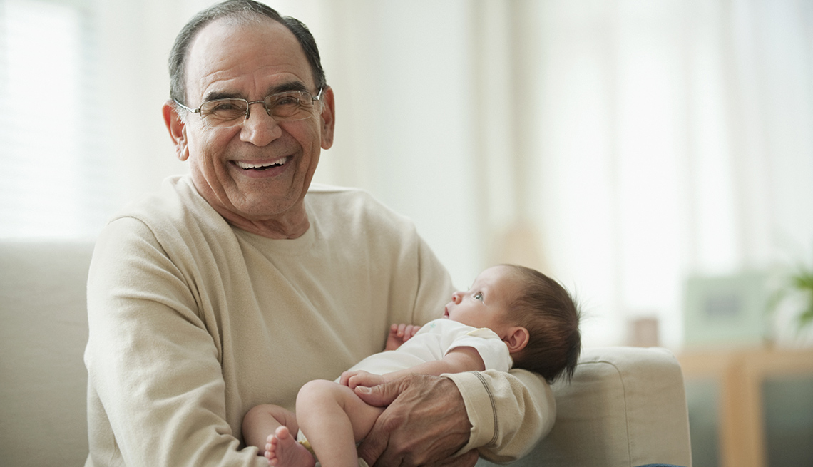 Grandfather holds an infant baby while sitting and happily smiles into the camera