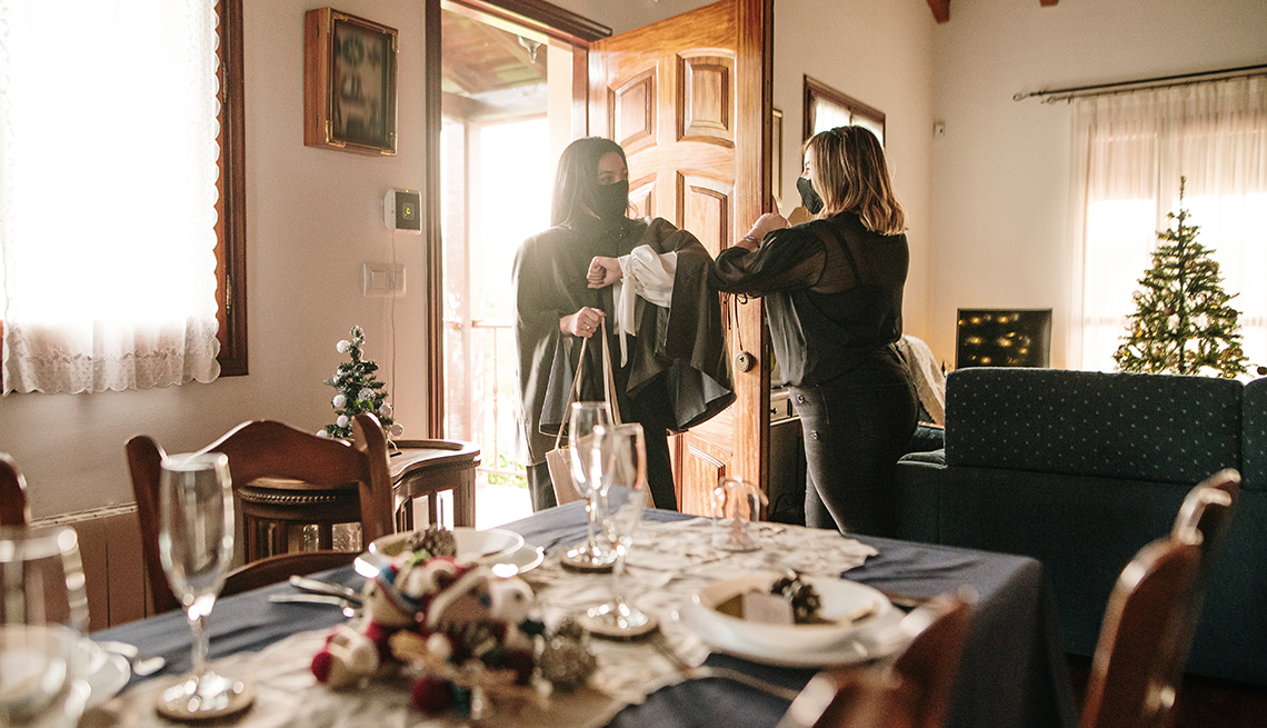 Dos mujeres se saludan en la entrada de una casa, con una mesa decorada de Navidad para cenar