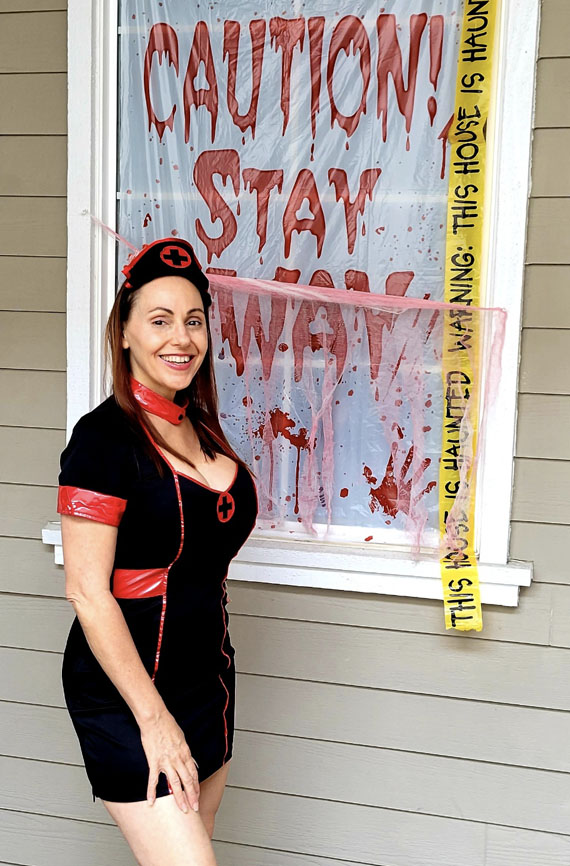 a photo of lynn ganter in a nurse costume in front of a blood splattered window with caution tape that says caution stay away and warning this house is haunted