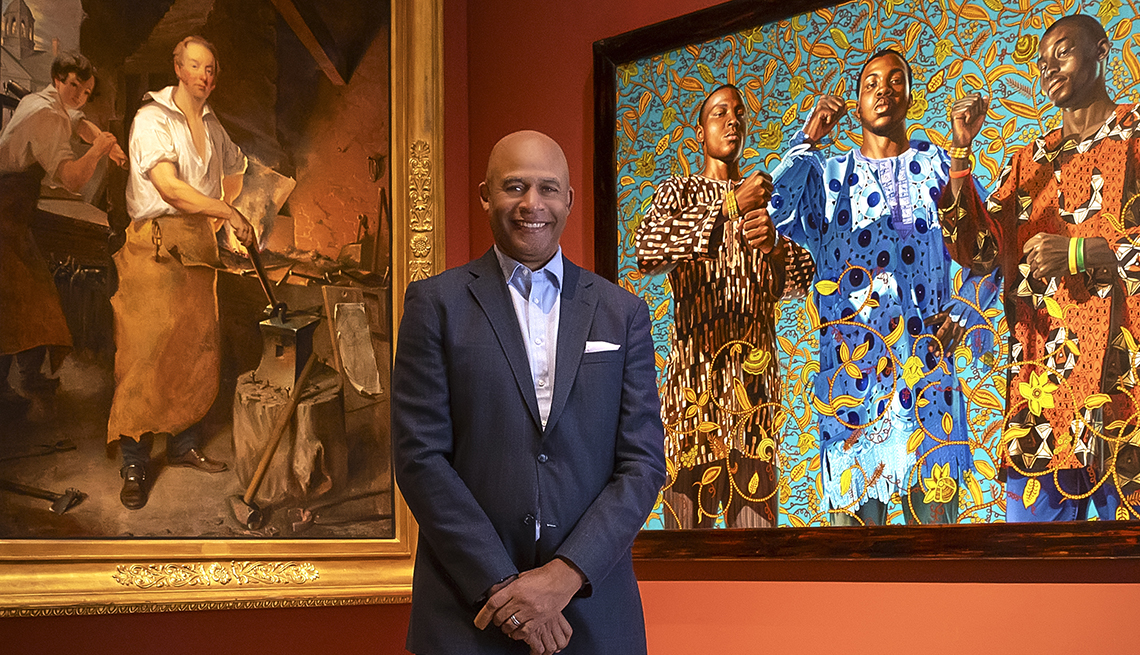eric pryor the president of the pennsylvania academy of fine arts tands in front of two paintings on the left is pat lyon at the forge by john neagle and on the right is three wise men greeting entry into lagos by kehinda wiley