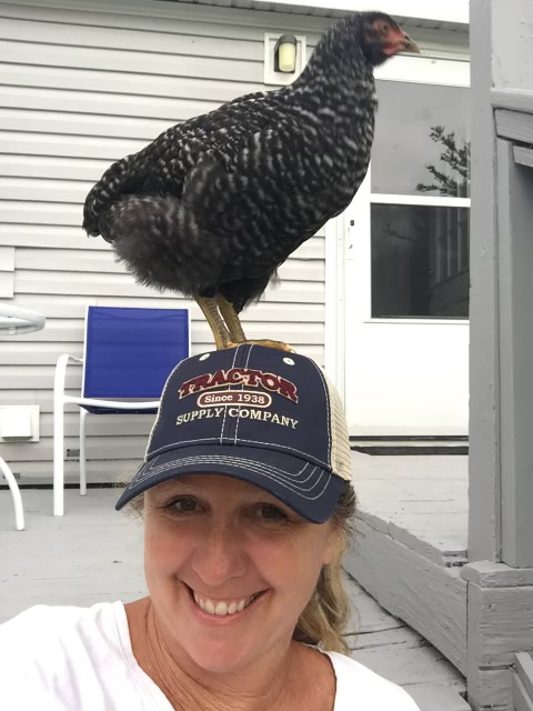 a smiling woman in a baseball hat with a chicken standing on her head