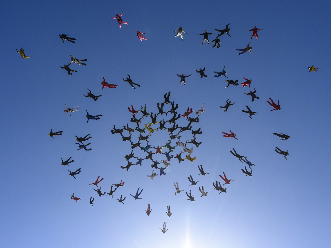 group of over fifty people skydiving and creating a linked formation as seen from below against the blue sky