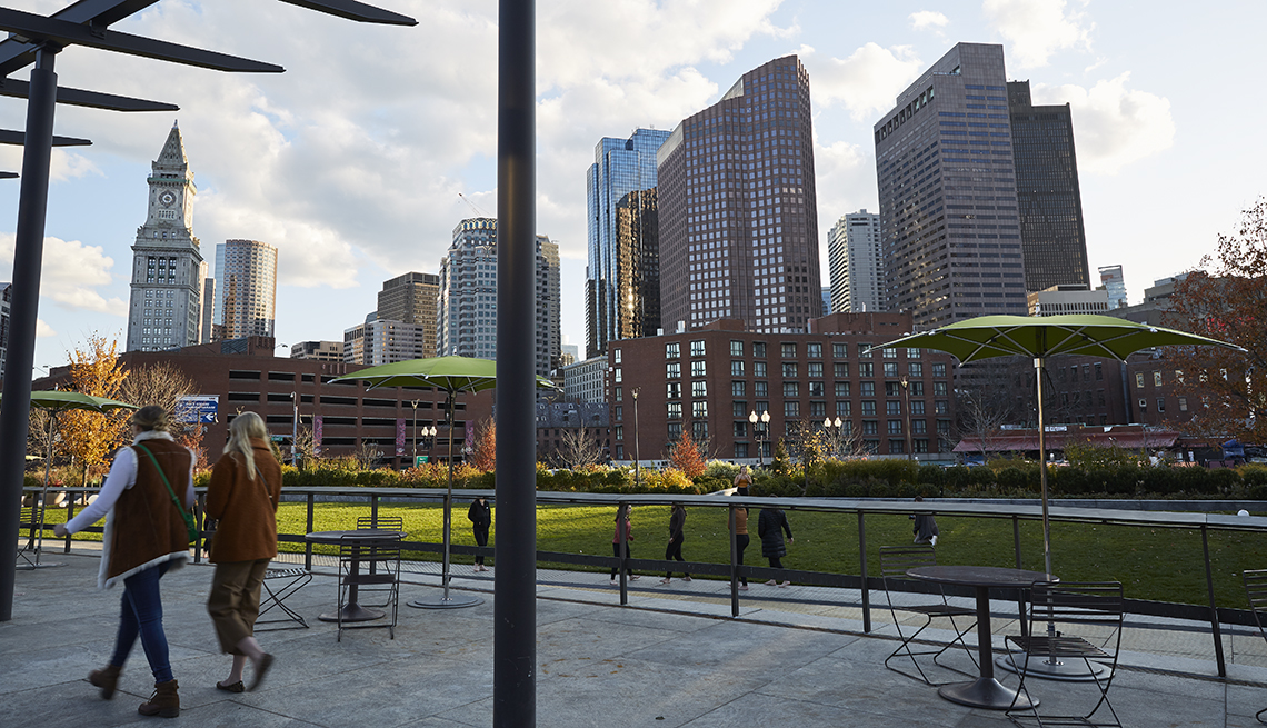two people walk along the greenway park area in boston with the city skyline rising up above them across a lawn