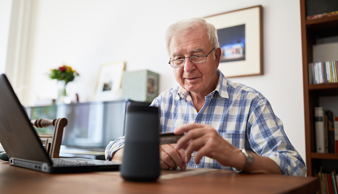 older man sitting at desk in home office holding a credit card and talking to smart speaker while making an online purchase