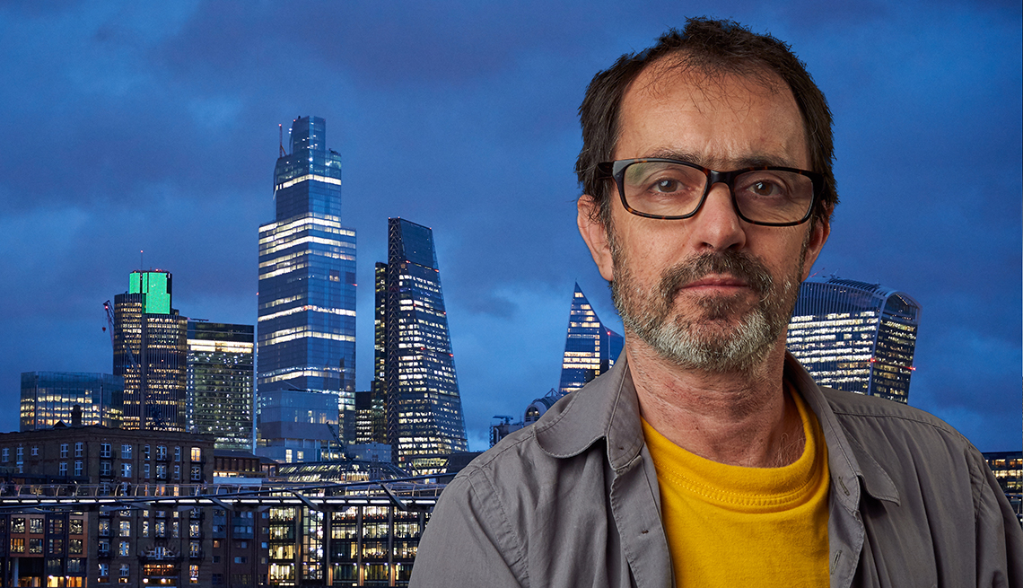 Man wearing glases sits in front of obviously fake tv like background.Wearing a grey shirt and yellow T-shirt. skyscrapers and blue sky