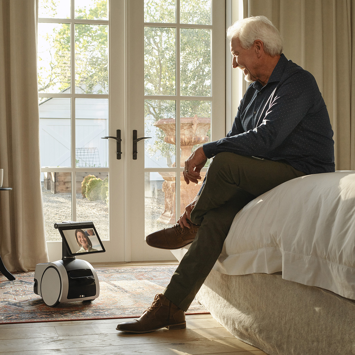 El robot Astro de Amazon interactúa desde el piso de una habitación con un hombre sentado en la cama
